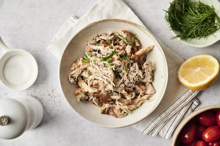 A bowl of shredded Greek chicken, garnished with fresh herbs. Surrounded by lemon, dill, cherry tomatoes, and salt, creating a fresh Mediterranean feel.