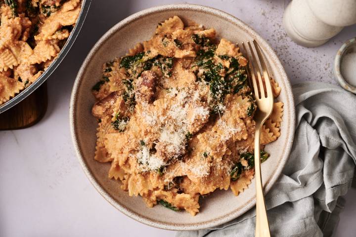 Easy High Protein Pasta Creamy high-protein pasta made with bowtie noodles, sausage, spinach, and ricotta, served in a beige bowl and topped with grated Parmesan cheese. A gold fork rests on the side.