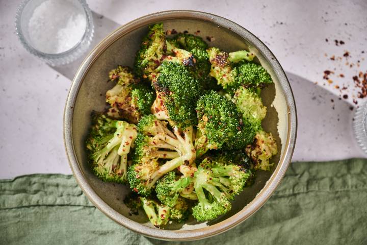 A bowl of roasted broccoli with crispy edges, sprinkled with red pepper flakes, sits on a light surface beside a salt dish.