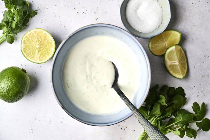 A bowl of creamy lime crema with a spoon, surrounded by fresh cilantro, lime halves, and a dish of sugar.