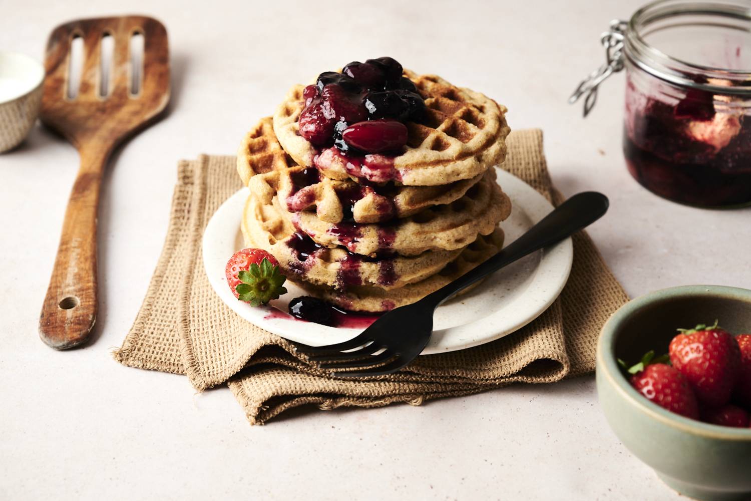 Stack of golden low carb waffles topped with warm berry compote and syrup, served on a small plate with a black fork, fresh strawberries on the side, and a rustic jar of fruit sauce in the background.