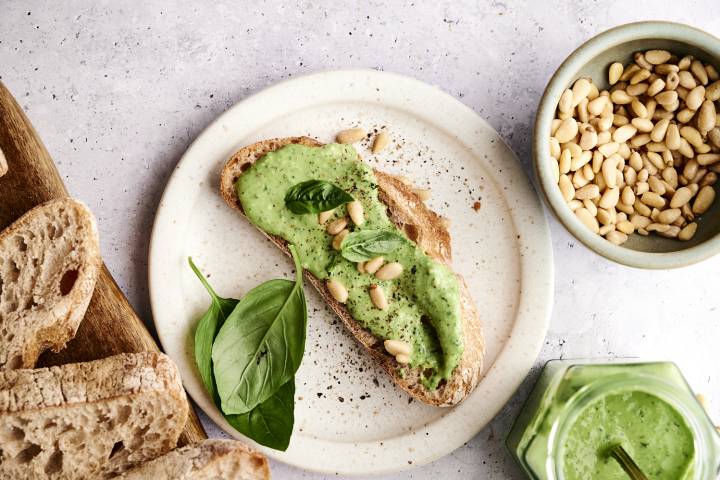 Skinny Pesto A slice of whole grain bread topped with green basil pesto, pine nuts, and fresh basil leaves, beside a bowl of pine nuts.