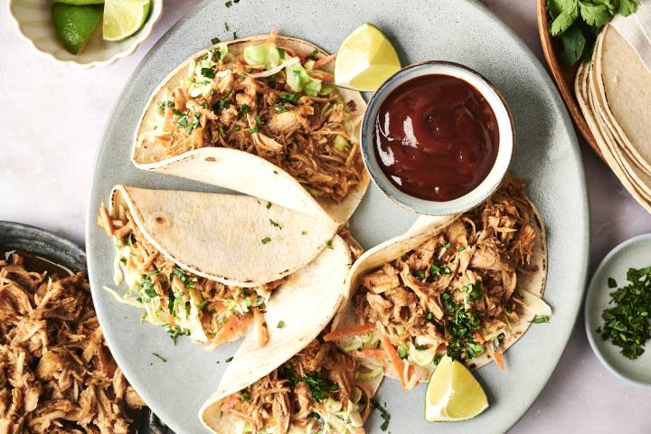 Three tacos filled with shredded meat and slaw, garnished with cilantro and lime, served with a bowl of sauce on a gray plate.