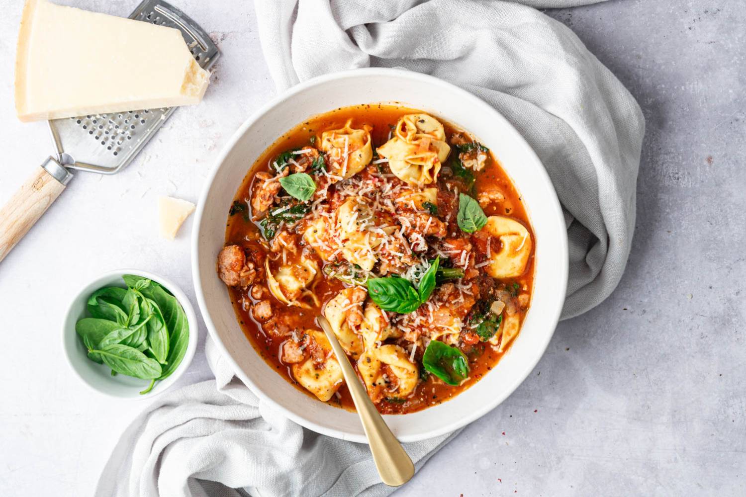A warm and hearty bowl of Slow Cooker Tortellini Soup filled with tender tortellini, flavorful sausage, spinach, and tomatoes, topped with freshly grated Parmesan and basil for a comforting finish.
