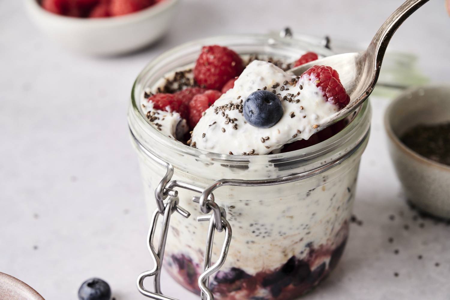 Spoon scooping creamy yogurt chia seed pudding topped with raspberries, blueberries, and chia seeds from a glass jar layered with fruit at the bottom.
